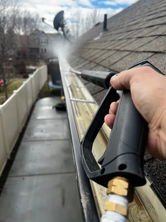 Reference photograph of a worker rinsing a residential gutter beside a shingle roof.